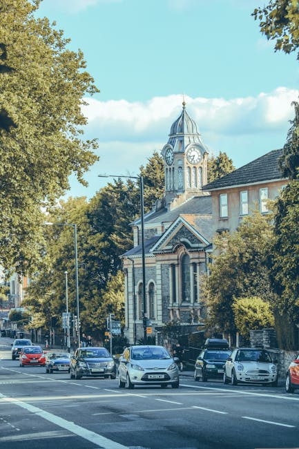 A wide street scene in Chiswick featuring a row of parked cars along the pavement, with a church building that has a prominent clock tower and a dome at the top visible in the background. The porch of the church has arched windows and decorative brickwork, with mature trees lining the street providing greenery. In the foreground, a loading area shows a variety of cardboard boxes, wrapped furniture, and packing materials being prepared for home relocation. A moving trolley and straps are visible near the boxes, indicating ongoing furniture transport and packing activities carried out by movers like Movers Chiswick. The lighting is natural, with clear skies and sunlight casting soft shadows, illustrating a typical house removals setting during a moving process in a residential urban area close to Bedford Park. This scene captures the logistics involved in local removals, packing, and furniture transportation, reflecting the services provided by Movers Chiswick.
