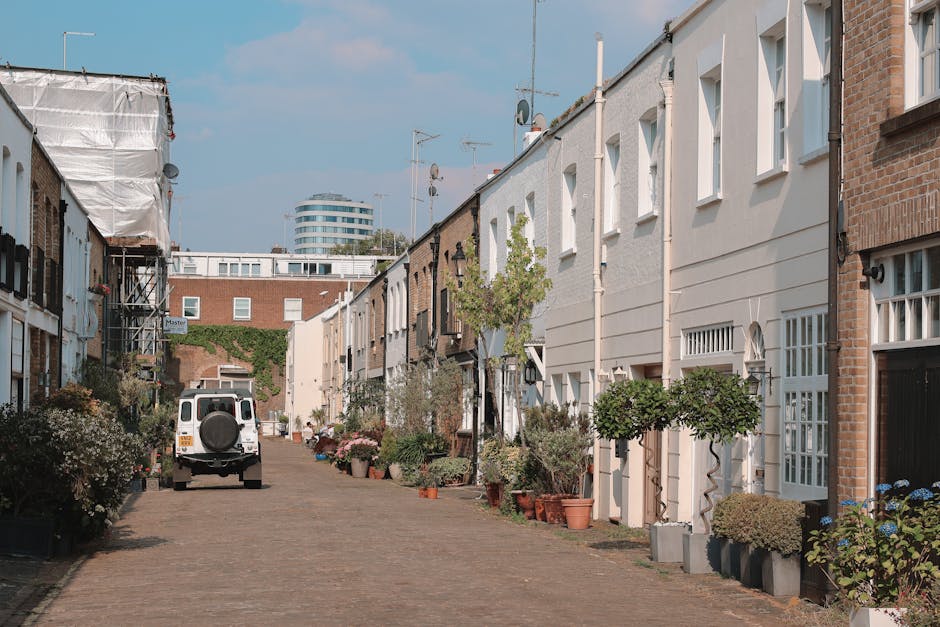 A narrow residential street in Bedford Park, Chiswick, with white and brick terraced houses on both sides. The street features a cobblestone or brick paving, with groupings of potted plants and small trees placed along the front of the properties, some housed in large ceramic or plastic pots. Towards the left side of the image, a white van with a spare tyre mounted on the rear is parked on the street, likely involved in a house removal or moving process. The scene is well-lit with natural daylight, and the background includes a modern, multi-storey building with curved architecture, contrasting with the traditional housing. This setting illustrates typical logistics involved in packing, loading, and transporting furniture and boxes during a home relocation, with the presence of a moving vehicle and prepared property exterior captured for content regarding house removals in Chiswick, supported by Movers Chiswick.