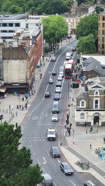 An aerial view of Chiswick High Road in London, showing a two-lane street with several cars and vans parked along the curbside, as well as some vehicles in motion, with pedestrians walking on the sidewalks. On the right side, a small white building with a pitched roof is visible, while on the left side, a red brick building with multiple floors houses shops and businesses, including La Perla, with storefront signage. Tall, green trees line the sidewalks and partially obscure the upper floors of surrounding buildings, creating a mix of urban and natural environments. A moving van or similar vehicle may be present as part of a home relocation or furniture transport process. The scene captures typical city street activity and can relate to house removals or moving services, with visible urban infrastructure, parking arrangements, and pedestrian activity, consistent with the context of house removals in the Chiswick W4 area. The image aligns with the services offered by Movers Chiswick and the house removals theme.
