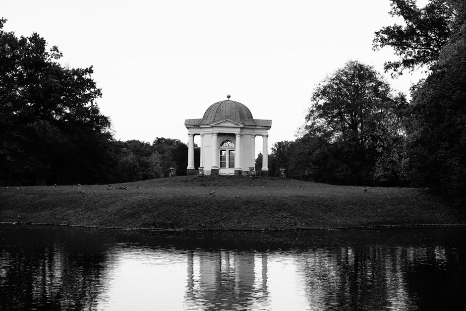 A scenic view of a tranquil pond with numerous green lily pads floating on the surface, surrounded by lush green trees with dense foliage on both sides. In the background, a decorative pavilion with a