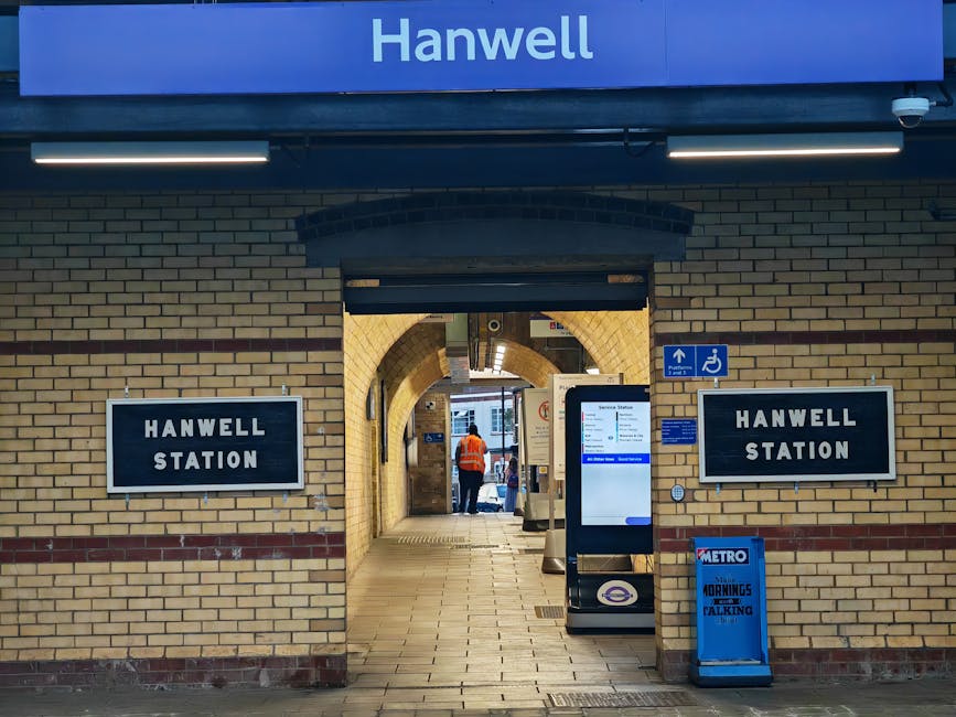 View of the entrance to Hanwell Station in Chiswick with a brick façade, featuring two black and white station signs on either side of the archway. Inside the station, a person in an orange high-visibility vest is visible, standing near the ticket machines and digital information screens. The platform area is illuminated by overhead lighting, and the corridor leads toward the platform and outdoor area. In front of the entrance, there are various objects associated with home relocation and furniture transport, including a digital display kiosk, a blue Metro newspaper stand, and a small blue sign indicating accessible facilities. The scene captures a typical station environment that Movers Chiswick might navigate during a house removal, highlighting the importance of careful planning for loading and unloading furniture or boxes as part of packing and moving logistics.