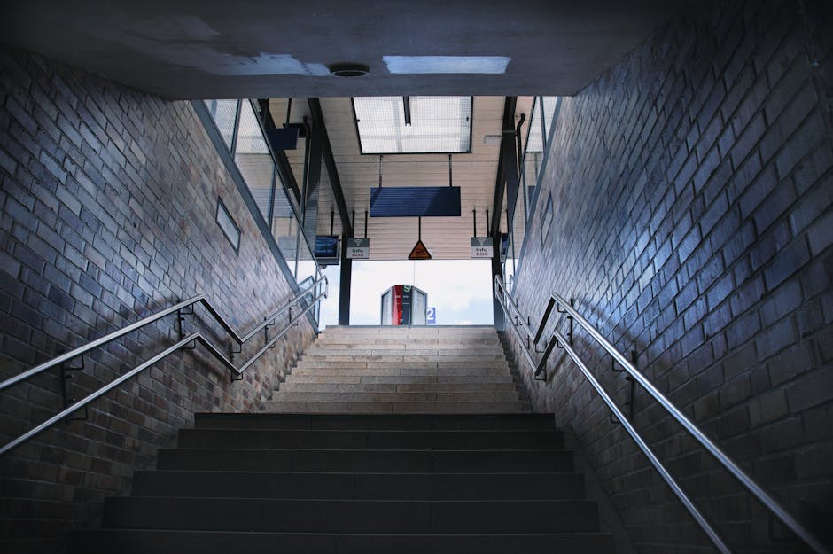 Interior view of a staircase leading up to Turnham Green station entrance, with brick walls on either side and metal handrails on both sides. The steps are made of dark material, with a section of lighter steps at the top near the doorway. Natural light illuminates the upper part of the stairs through a glass ceiling, revealing signage and the station entrance ahead. The scene captures part of a home relocation process, with the setting indicating a transfer point for furniture transport managed by Movers Chiswick, related to house removals in the Chiswick area. The overall environment is clean and structured, suitable for loading and unloading activities involved in packing and moving services.