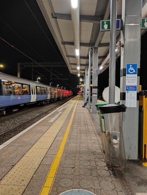 View of the entrance to Hanwell Station in Chiswick with a brick façade, featuring two black and white station signs on either side of the archway. Inside the station, a person in an orange high-visibility vest is visible, standing near the ticket machines and digital information screens. The platform area is illuminated by overhead lighting, and the corridor leads toward the platform and outdoor area. In front of the entrance, there are various objects associated with home relocation and furniture transport, including a digital display kiosk, a blue Metro newspaper stand, and a small blue sign indicating accessible facilities. The scene captures a typical station environment that Movers Chiswick might navigate during a house removal, highlighting the importance of careful planning for loading and unloading furniture or boxes as part of packing and moving logistics.